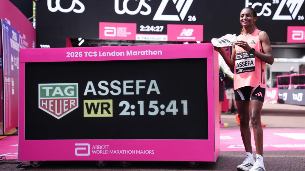 Ethiopia's Tigst Assefa stands on a pink podium holding a running shoe next to a large digital clock displaying her world record time of 2:15:41.