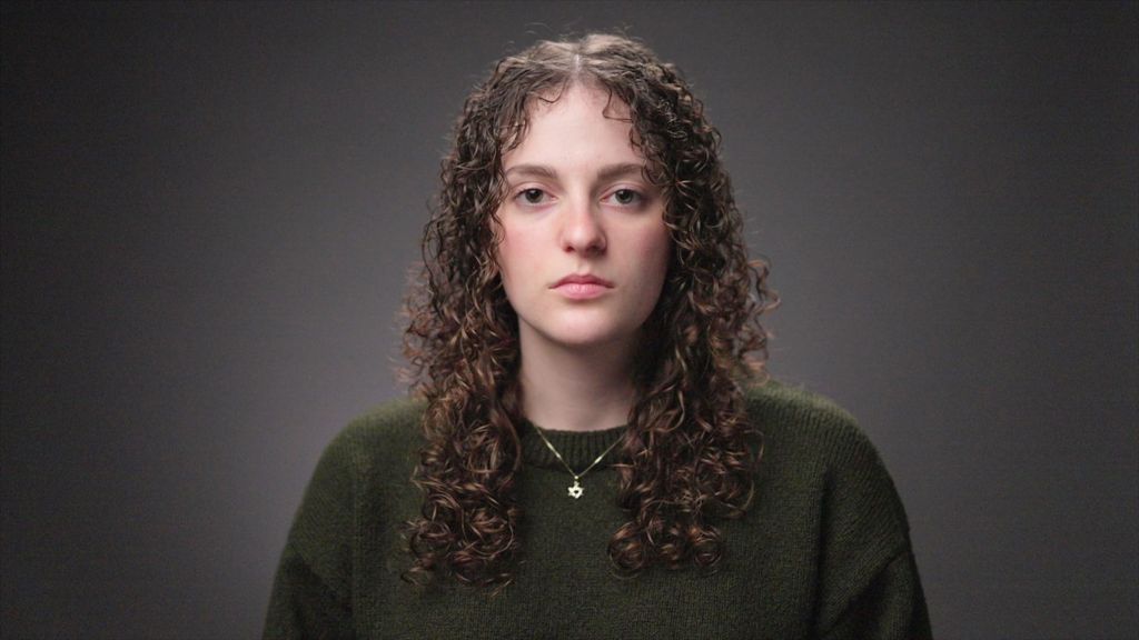 A woman looks at the camera. She has long curly hair and is wearing a dark green jumper and a Star of David on a chain around her neck