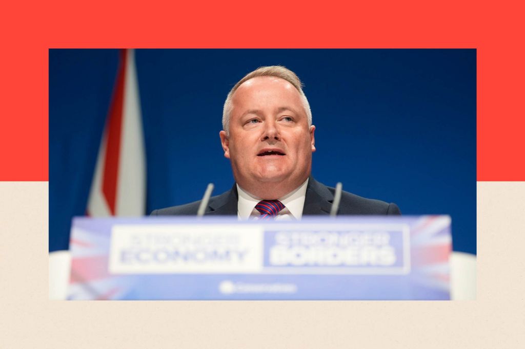 Darren Millar, the Conservatives' Senedd leader, stands in front of a blue backdrop and a union flag. In front of him is a lectern with a microphone. The words "STRONGER ECONOMY /STRONGER BORDERS" and the Conservatives logo appear on the front of the lectern. Mr Millar wears a dark suit with a white shirt and red and blue striped tie.