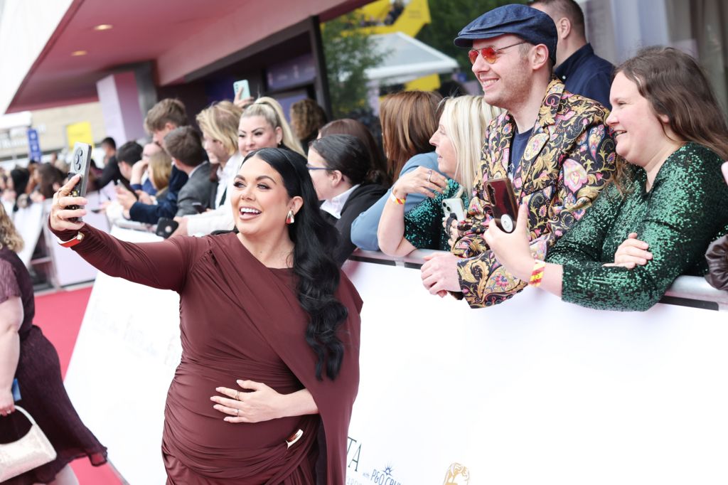 Scarlett Moffatt holding her stomach in one hand and a phone in the other, taking a selfie with some fans behind a barrier
