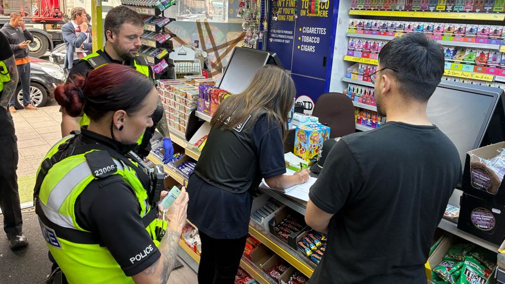 Police officers stand inside a convenience store as a shop worker and Trading standards official fill out paperwork at the counter, with shelves of sweets, cigarettes and colourful vaping products behind them.