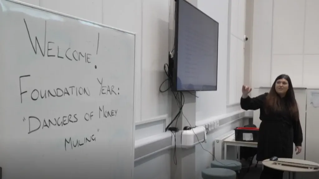 Criminology lecturer Dr Sam Mapston stood in a class room. She is pointing at a screen. Next to the screen is a white board with the words 'Welcome Foundation Year: Dangers of Money Muling' written on it. 