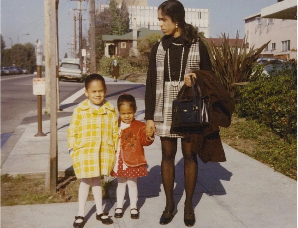 A little girl in a yellow coat, a little girl in a red coat and an Indian woman dressed in black and white stand on a sidewalk