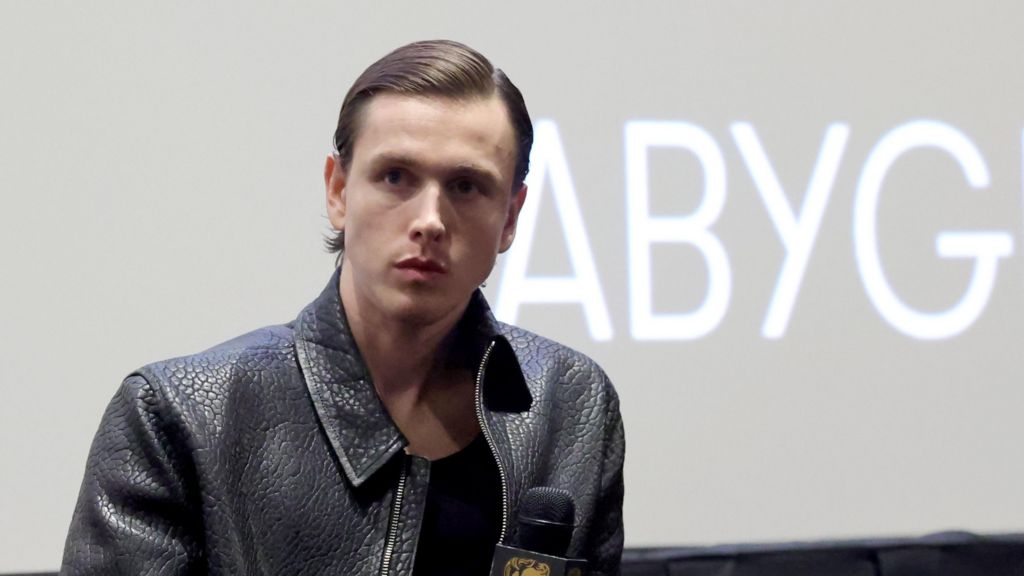 Harris Dickinson is dressed in a black leather jacket and holds a microphone