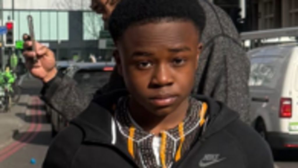 Abdul is pictured wearing a black hoodie and earphones looks towards the camera on a city street, with a car and a cyclist visible in the background.