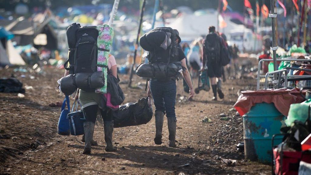 People wearing backpacks leaving the Glastonbury Festival site. They are walking alongside a row of bins