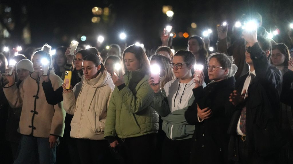 A group of women gather with their lights on their phones looking mournful.