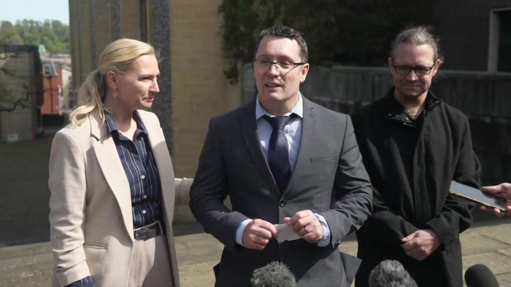 Christopher Trybus wearing a dark grey suit and navy tie, standing outside court delivering a statement to the press, with a folded piece of paper in his hands. A woman wearing a pinstripe shirt and light-coloured suit stands beside him, holding onto his arm.