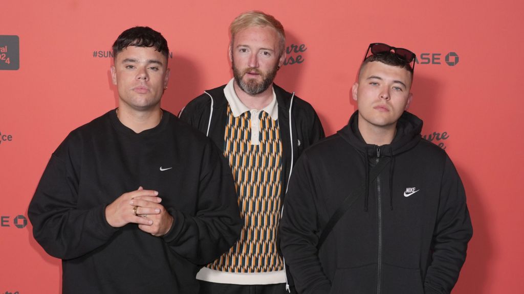 Kneecap members Moglai Bap, Mo Chara, DJ Provai standing together in front of a red wall.