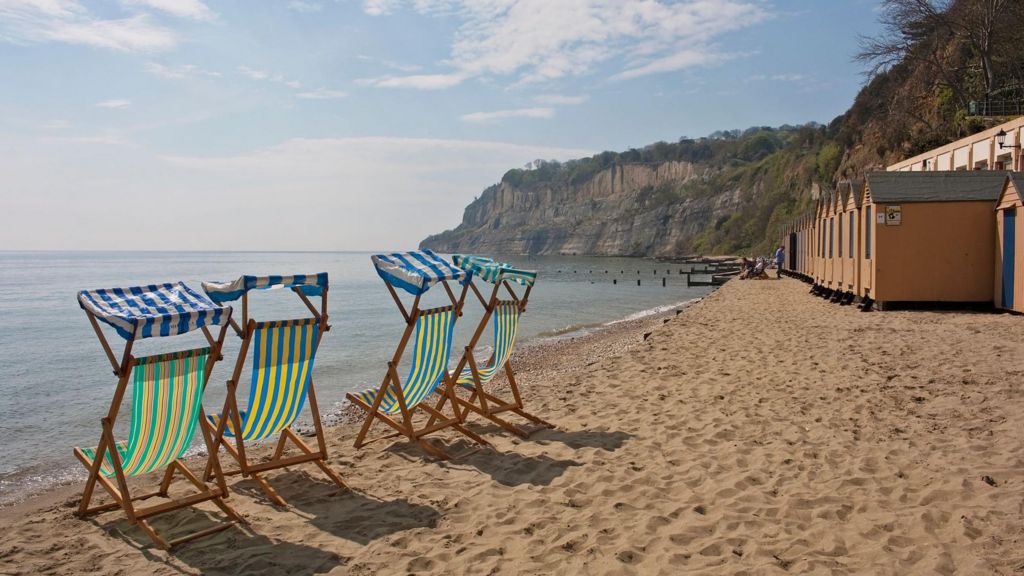 Brightly coloured striped deckchairs on the beach on the Isle of Wight. The sand is golden brown and the sea is light blue. There are wooden huts on the beach and cliffs in the background. The sky is clear and it is sunny.