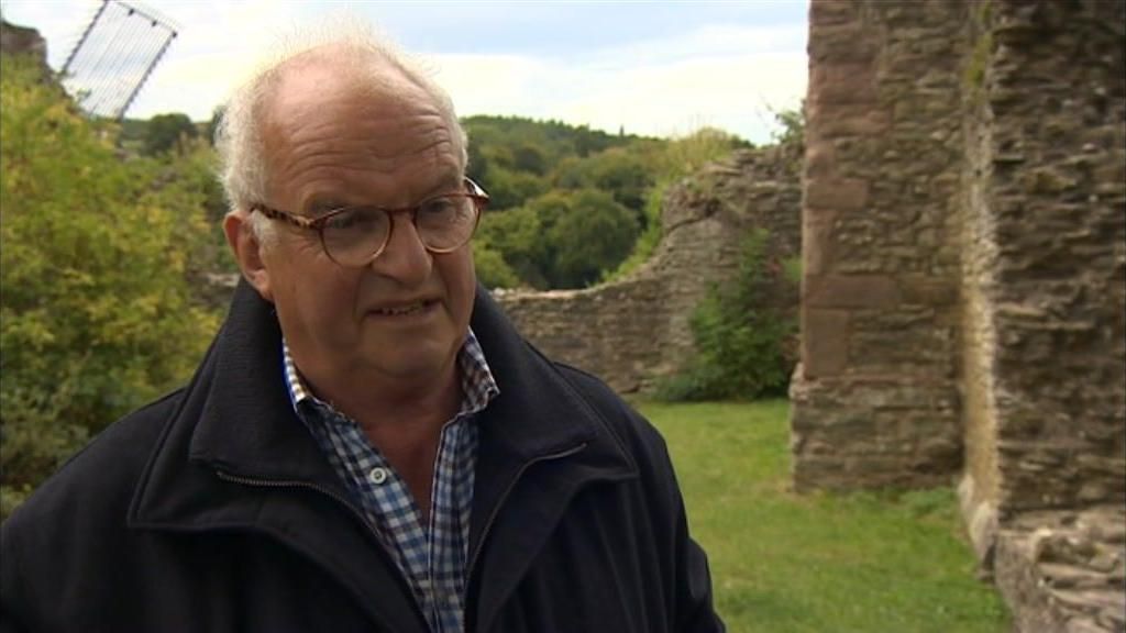 Colin Richards has white hair and glasses. He is standing next to a castle wall and some grass. He is wearing a dark blue jacket and a white and blue checkerboard shirt.