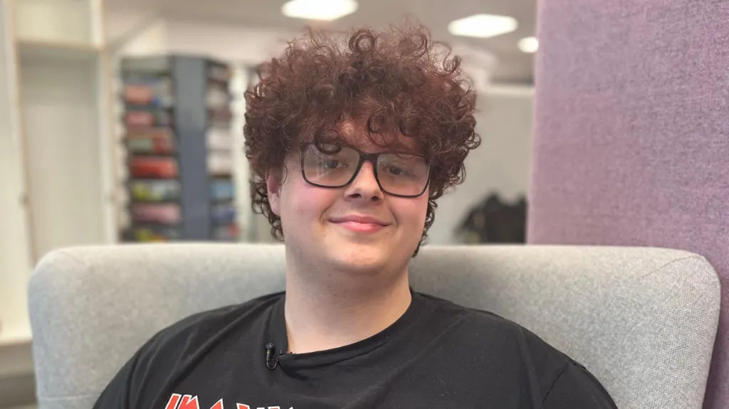 Cameron has hurly brown hair, glasses and is wearing a black t-shirt. He is sitting in a  grey high-backed armchair in a library and looking directly into the camera.  
