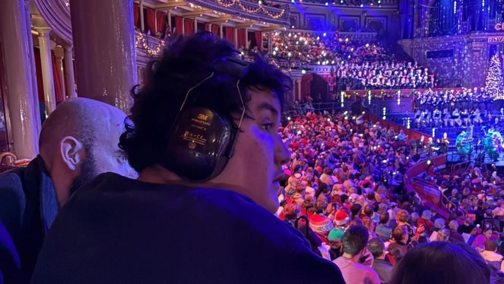 A boy with large headphones on looks across the crowd at the Royal Albert Hall. There are festive lights and lots of santa hats in the background.