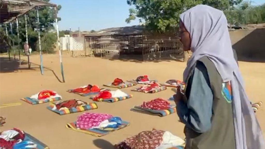 Piles of colourful blankets are arranged in rows on the ground. Hafiza is in the foreground wearing a grey robe and light purple headscarf.