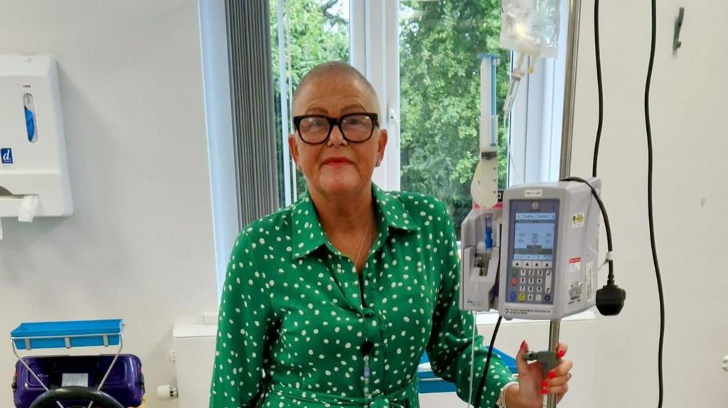 Tracie having chemotherapy treatment, she wears a green shirt with white dots on it and has glasses on. Her head is shaved and she holds a pole in her left hand which has medical equipment on it