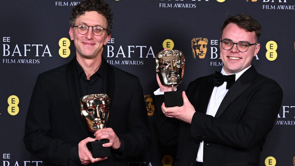 David Borenstein on the left. Dark curly hair, glasses, smiling. Black suit.
and Pavel Talankin on the right. brown hair, glasses, wide smile. Black dinner jacket and bow tie.
Both holding the Documentary Award for 'Mr Nobody Against Putin' during the BAFTA Film Awards in February.
Background is black with BAFTA logo and words EE BAFTA Film Awards