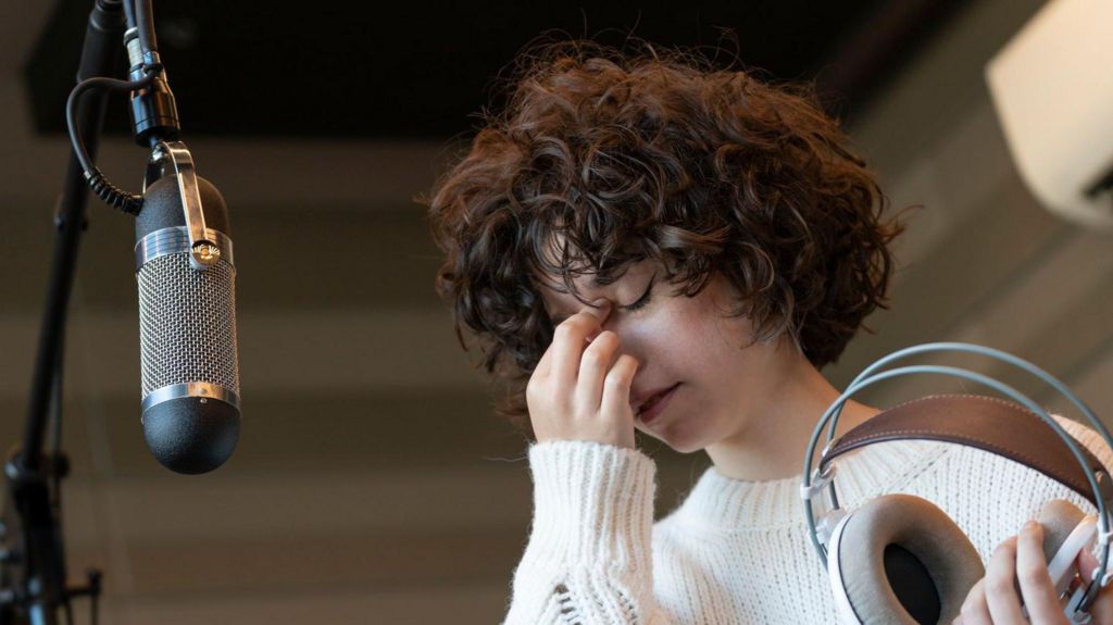 A stock image of a female vocal artist hand covering her face, her eyes closed, standing in front of a large microphone on a stand