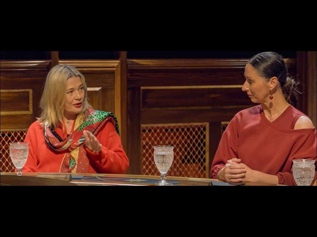 Harriet, in a red cardigan and red scarf, is sat next to Rachel, in a red dress, at the Traitors round table
