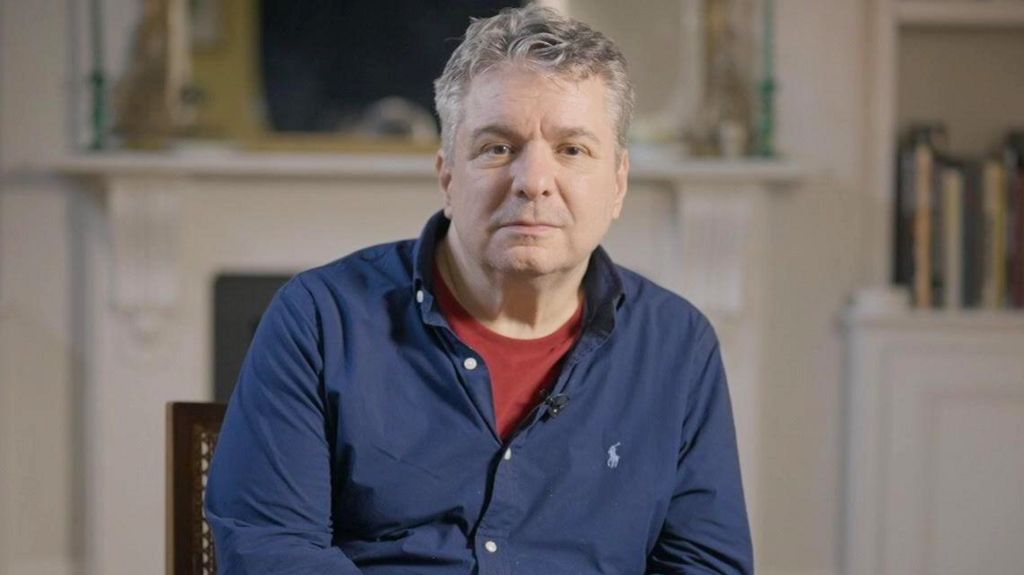 Dr Ittai Gradel looking to camera in the centre of the image. In blue shirt and maroon top underneath. Behind him books and fire place