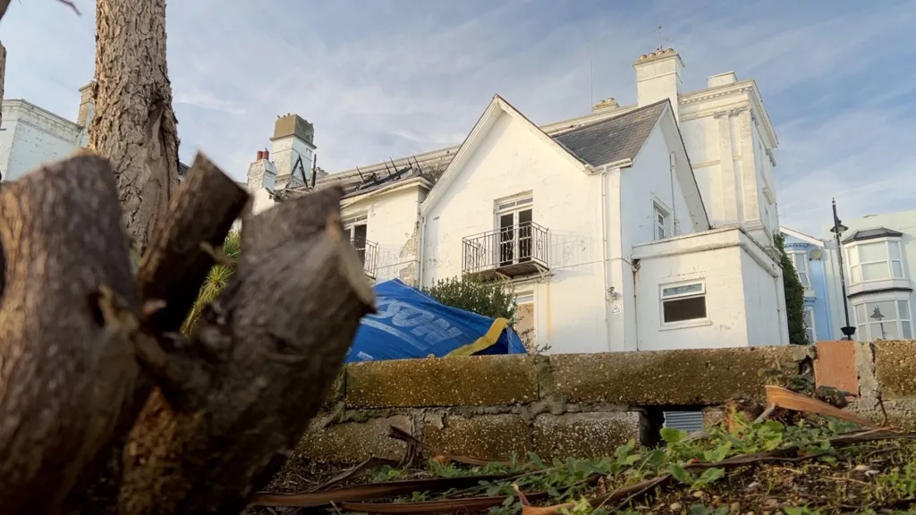A white hotel in the background is in focus. Part of its roof is burned out and the windows are smashed. A blurry tree stump and a blue bag full of construction materials is in the foreground.