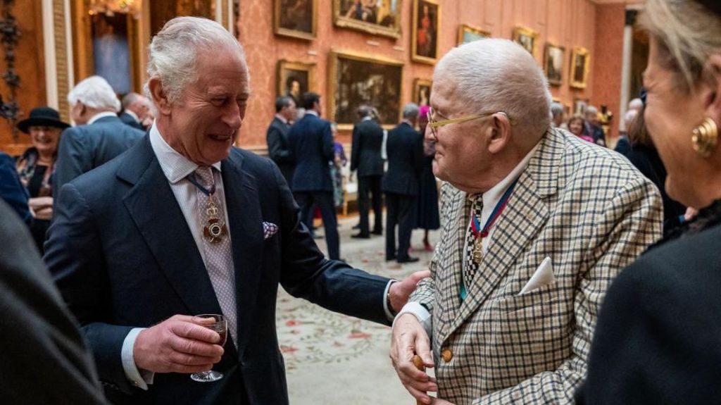 King Charles III talks with Mr David Hockney during a luncheon for Members of the Order of Merit at Buckingham Palace on November 24, 2022 in London, England. The King has his hand on the arm of Hockney. David Hockney is wearing a distinctive suit with the two men smiling at each other.
