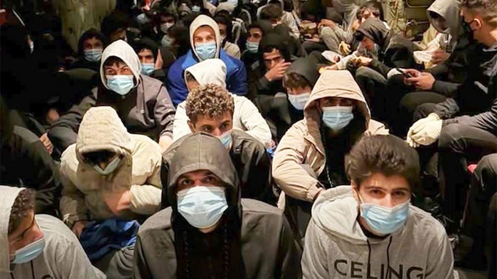 A large group of young men wearing hooded sweatshirts and light-blue surgical masks sit closely together on the floor in a crowded indoor space. Most face forward toward the camera, with some looking downward or to the side. The setting appears dimly lit, with people packed tightly in rows, filling the frame from foreground to background. Clothing is mostly dark or neutral tones, and the confined arrangement suggests limited space.
