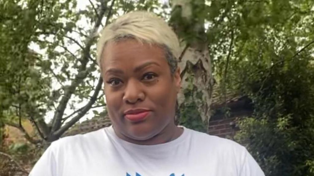 Shantel Morris wears a white t-shirt and has short bleached blonde hair in a crew cut. In the background is a brick wall and trees. 
