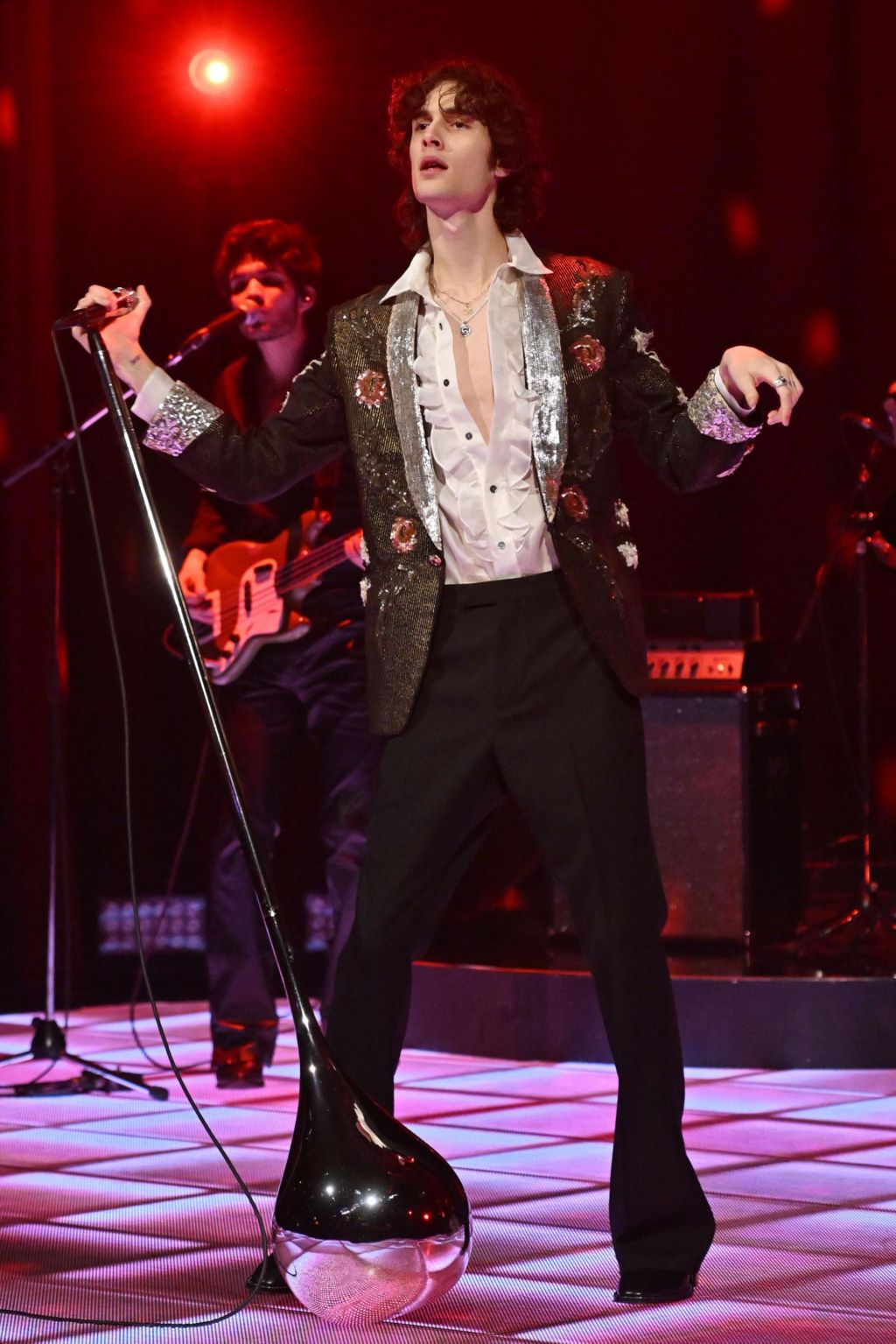 Sombr in a half-unbuttoned white shirt and decoratively emroidered black jacket, with one hand on a leaning microphone stand, while performing on The Graham Norton Show in November 2025.