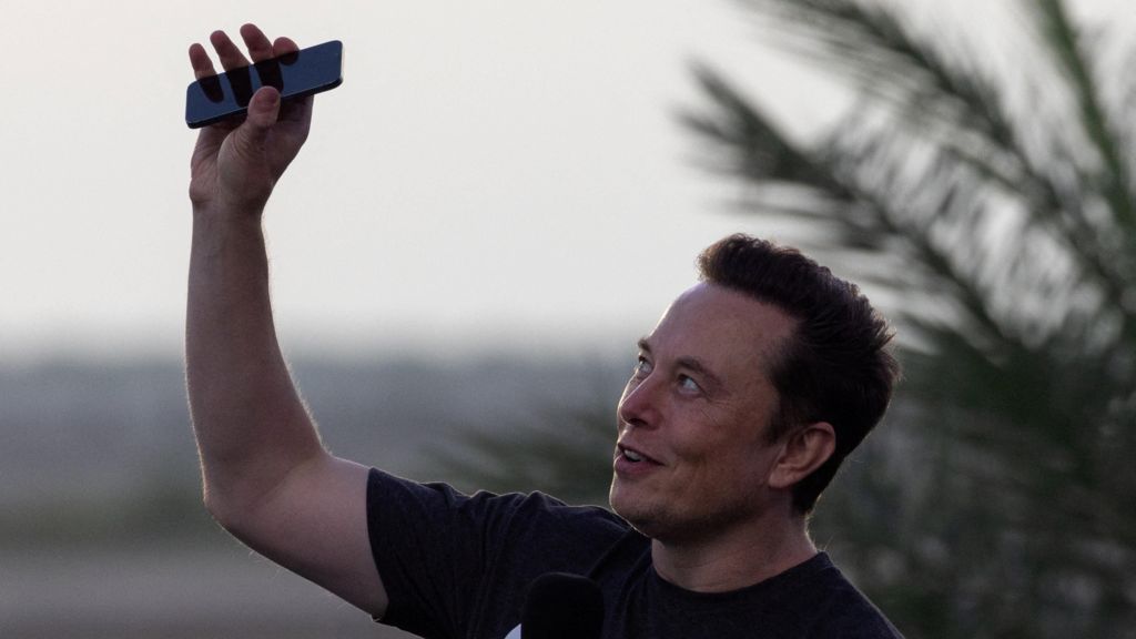 Elon Musk wearing a black T-shirt and holding his phone above his head.