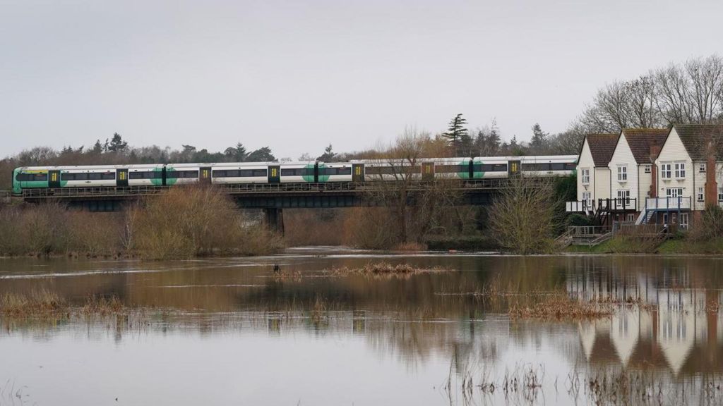 A green, white and yellow train crosses a bridge above a flooded river with three white houses on the right side riverbank over a