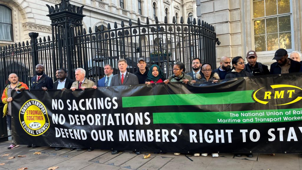 RMT general secretary Eddie Dempsey (centre) with workers holding a large black and green banner which reads "NO SACKINGS, NO DEPORTATIONS, DEFEND OUR MEMBERS' RIGHT TO STAY"
