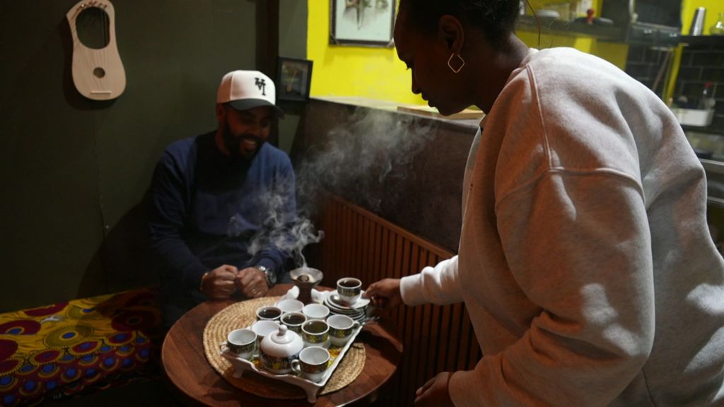 A woman serves a tray of steaming tea to a man who sits smiling.