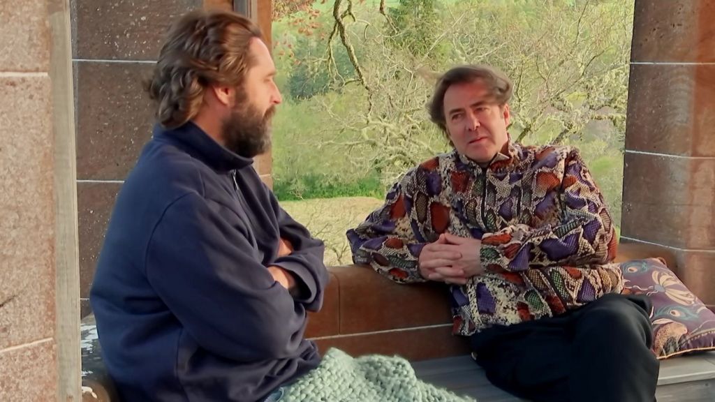 Jonathan Ross wears a patterned sweaters talking to another with his arms crossed