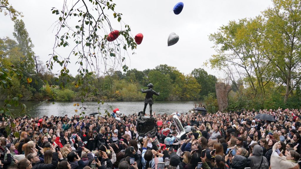 A large crowd gather round a statue, releasing love heart-shaped balloons