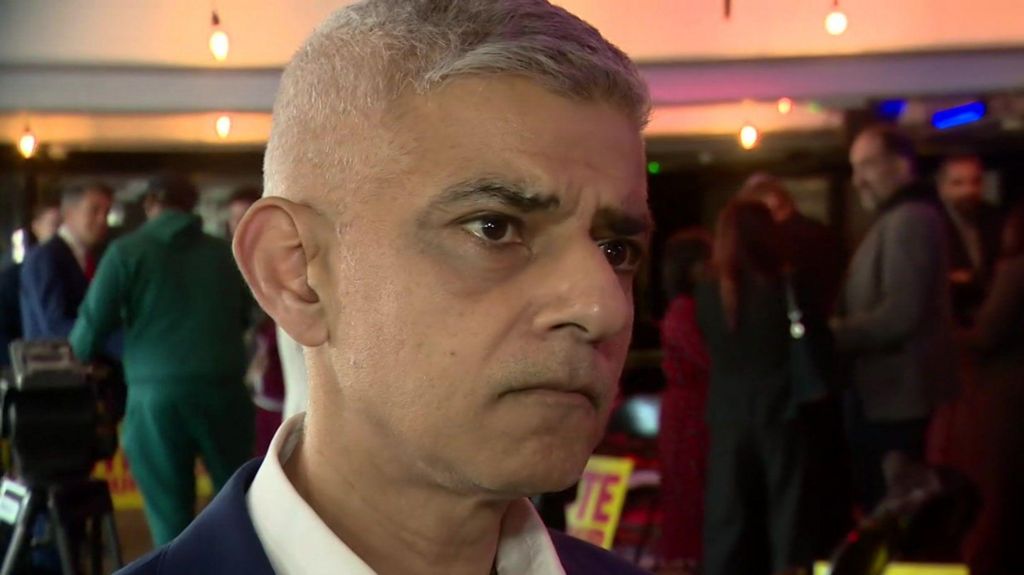 A close up image of Sir Sadiq Khan. He is wearing a dark blue suit jacket and an open-necked white shirt. Behind him are a crowd of people in an event space. 