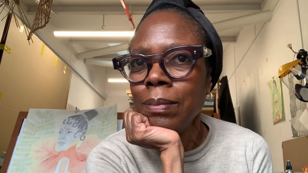 A woman wearing glasses looks at the camera. She has her chin in her hand and behind her is a painting of a woman wearing an orange dress with a green background. 