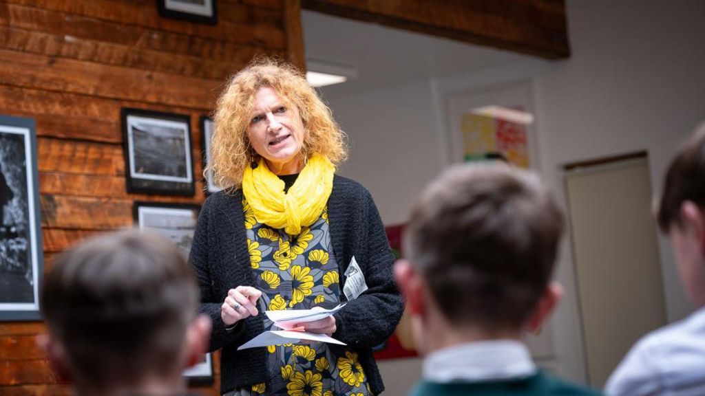 A woman is speaking to a group of school children. She has ginger hair and is wearing a yellow scarf.