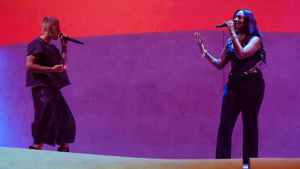 Justin Bieber and Tems pictured on stage at Coachella, in front of a red and purple background.