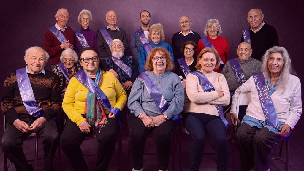 17 members of the choir stand and sit in formation, looking at the camera and wearing purple sashes. 