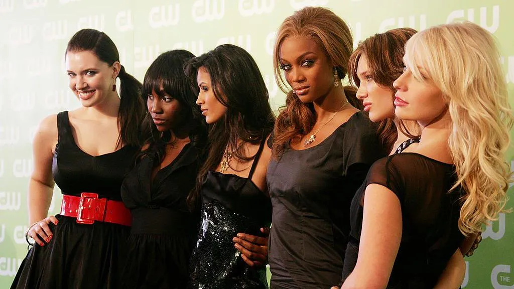 Tyra Banks looks at the camera as she poses with the cast members from America's Next Top Model. She is wearing a black top and has light brown hair swept to the side. 