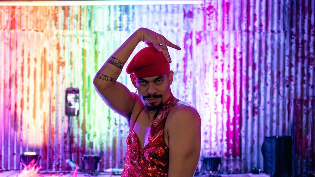 A man in a red sequin jumpsuit and beret pulls a pose