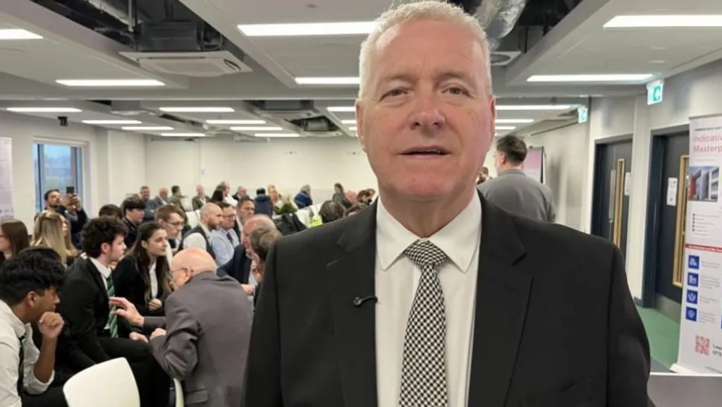 Head and shoulder shot of Ian Lavery with short, close cropped grey hair. He is wearing a dark jacket over a cream shirt and grey and white checked tie. Behind him are a number of people seated in a conference room.