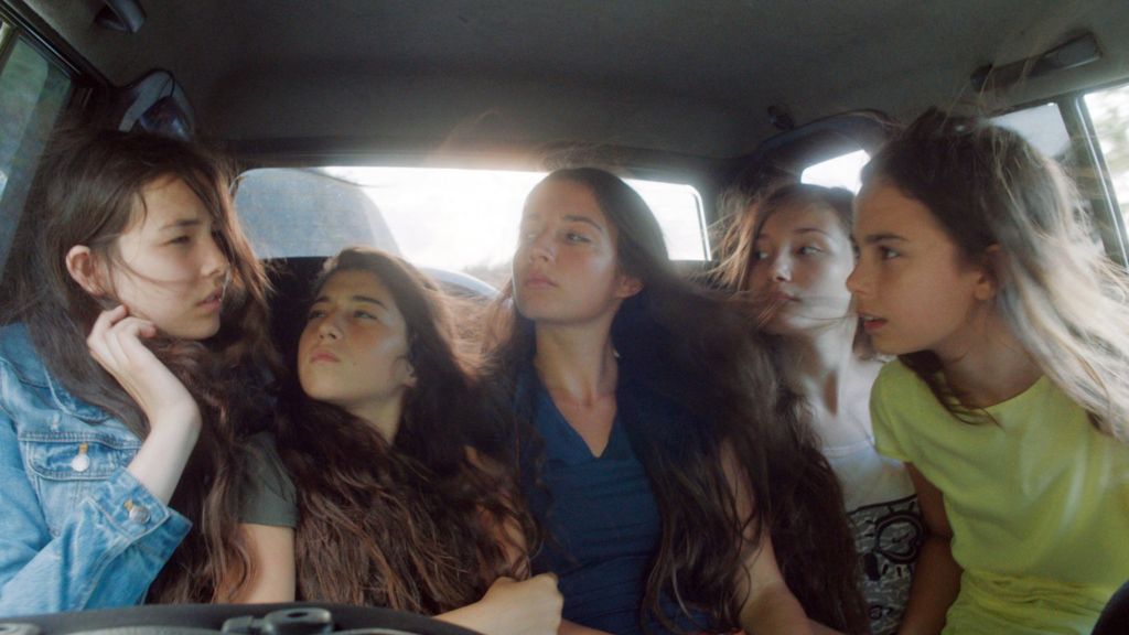 Image from the film Mustang. Five sisters talk to one another in the back seat of a car. Their hair is swept over their faces as they drive.