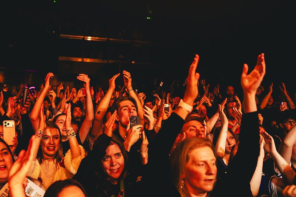 A view of the crowd during a Myles Smith show - the crowd's hands are in the air and people are smiling, while illuminated dark orange from the stage lights.