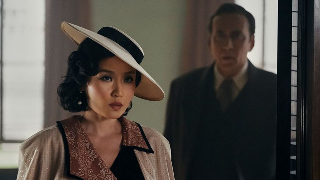 The character of Cat Hardy, a woman wearing a cream coloured hat and top with light red patterns. Behind her is the character of Bill Reilly, wearing a dark coat, tie and white shirt.