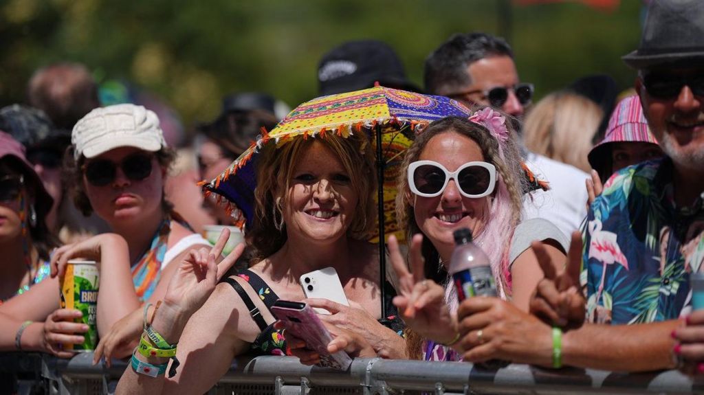 Two women standing under a sun umbrella at the Pyramid Stage at Worthy Farm with other festival-goers stood near them.