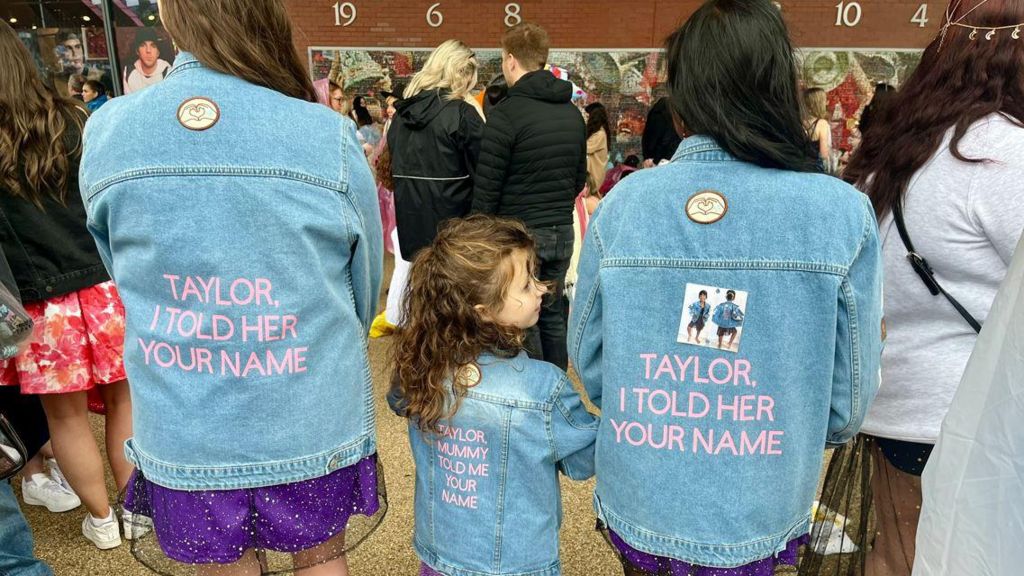 Lauren Stephenson, Florence, and Brinda Selvamanoharan facing their back to the camera to show their matching denim jackets with the words 'Taylor, I told her your name' in pink 
