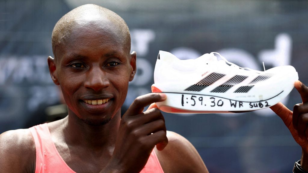 A close-up of Kenyan runner Sebastian Sawe smiling and holding up a white running shoe with "1:59.30 WR Sub2" handwritten on the sole to celebrate his record-breaking finish.