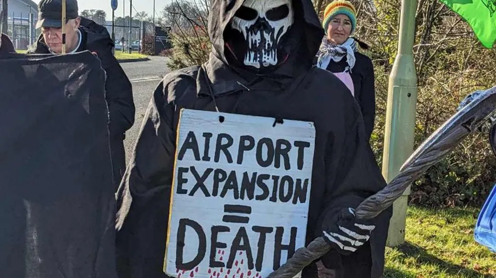 The image shows environmental protesters, including one person dressed as the Grim Reaper in a black hooded robe and skull mask, holding a white sign reading 