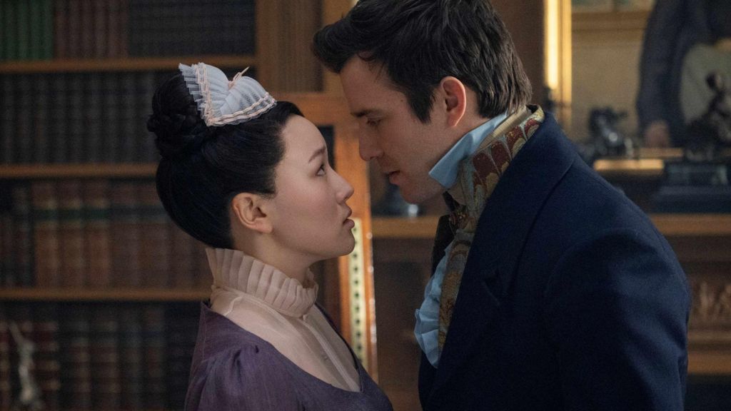 A woman and man stand facing each other. The woman (Sophie Baek) is in her maid's uniform, which is purple with white frills. The man (Benedict Bridgerton) is in a navy blue suit and shirt. They stand in front of a library and mirror